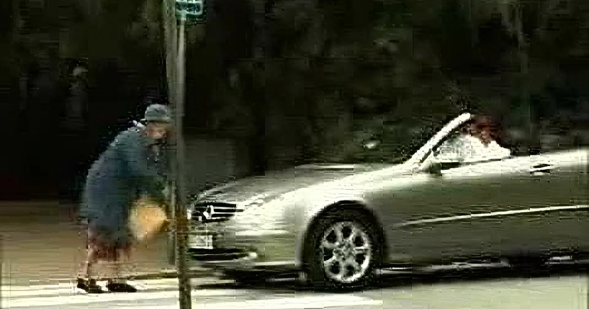 A person in a blue jacket and hat walks on a sidewalk near a pole and a silver car.