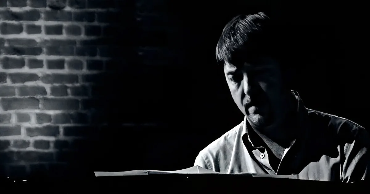A man in a dimly lit room, wearing a collared shirt, appears contemplative or focused.