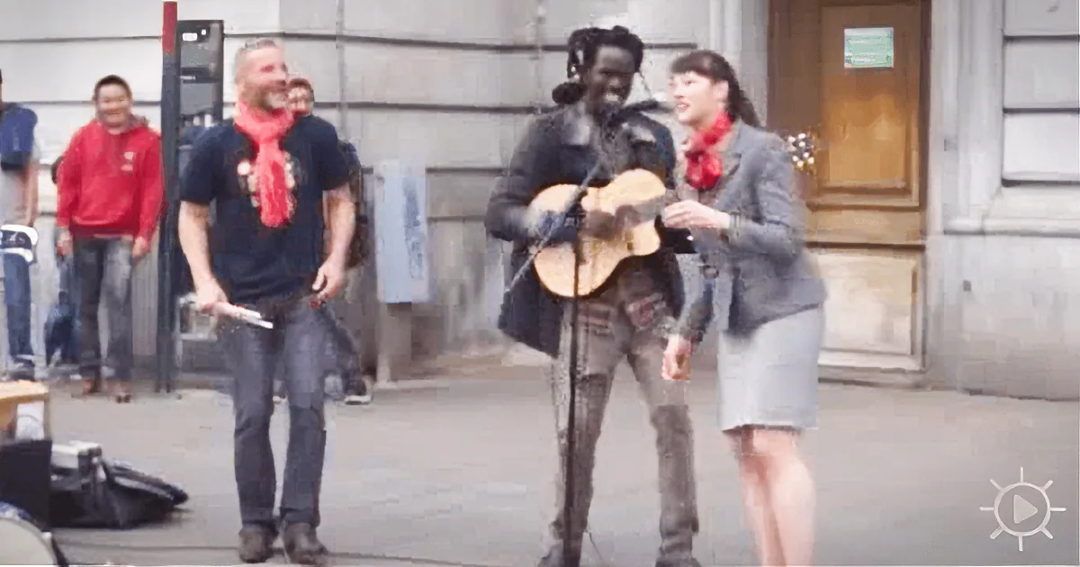 A man with dreadlocks plays guitar and sings on a street, accompanied by a woman, while a man in a red scarf laughs in...