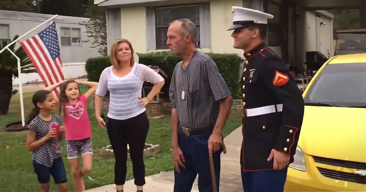 A Marine in uniform stands with a family in front of a house, holding an American flag.