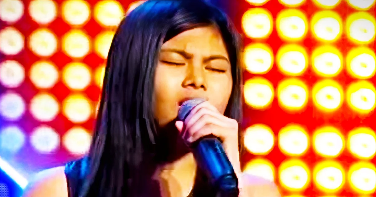 A person with long dark hair sings into a microphone against a vibrant, illuminated background.