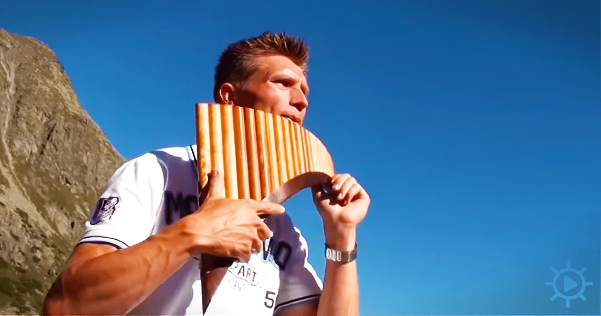 A man plays a wooden pan flute outdoors, with a clear blue sky and a mountain in the background.