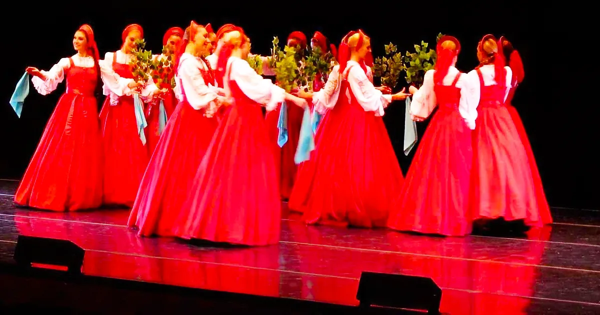 A group of dancers in red dresses performs on stage, holding bouquets and blue scarves.