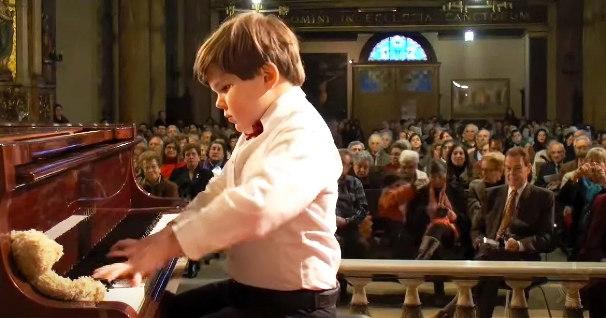 A young boy in a white shirt plays a piano in front of a large audience in a church.