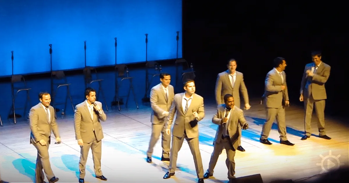 A group of men in suits perform on stage under blue lighting, holding microphones.