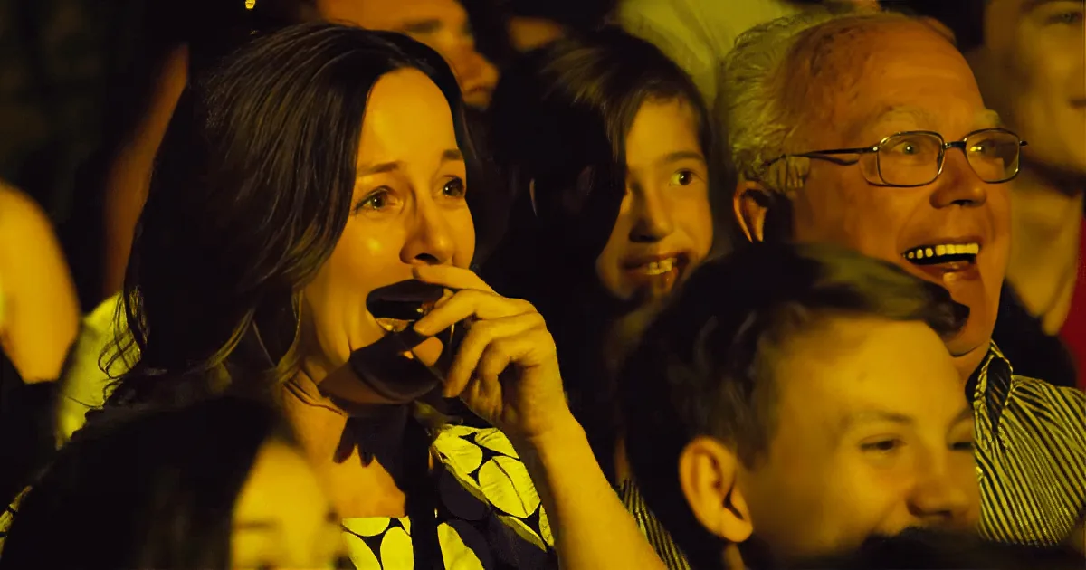 A woman in a yellow patterned shirt holds a microphone, smiling amidst a crowd of people.