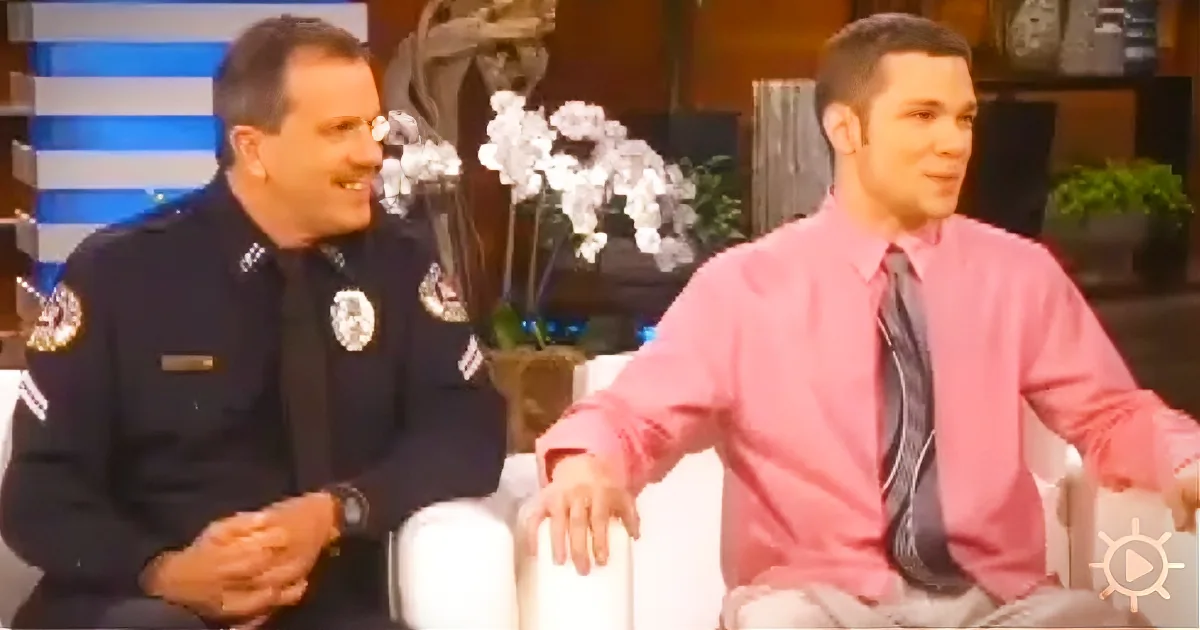 Two men, one in a police uniform and the other in a pink shirt, are seated and engaged in conversation.