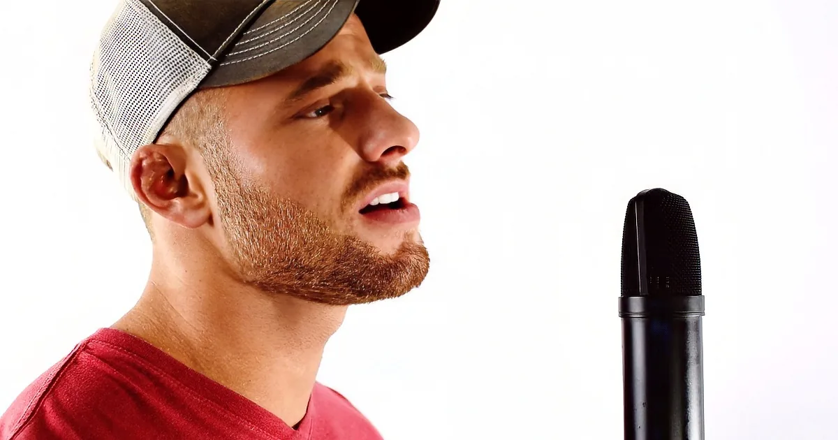 A man in a red shirt and cap sings into a microphone against a white background.