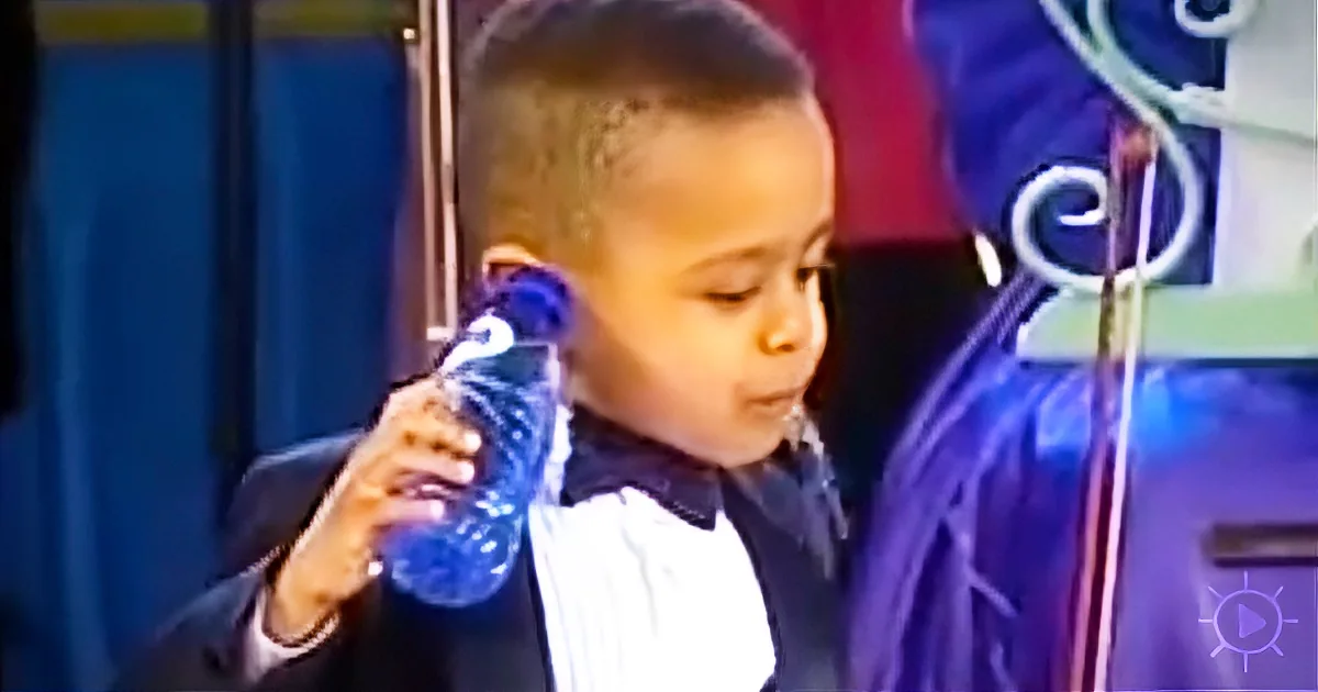 A young boy in a suit holds a blue water bottle, looking down.
