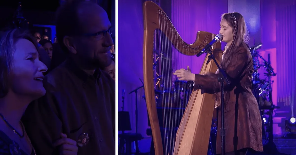 A woman plays a harp and sings into a microphone, surrounded by purple lighting and an audience.