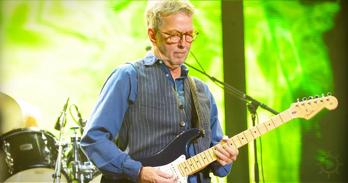 A man in glasses plays a Fender guitar on stage with a green backdrop.