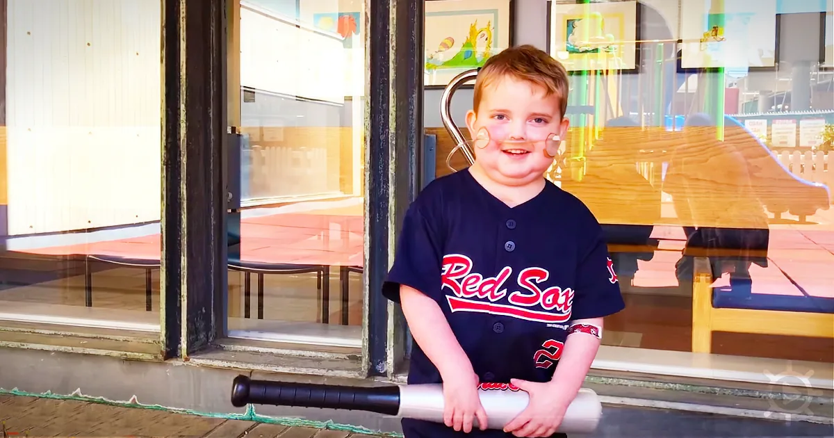 A young boy in a Red Sox jersey holds a baseball bat in front of a window.