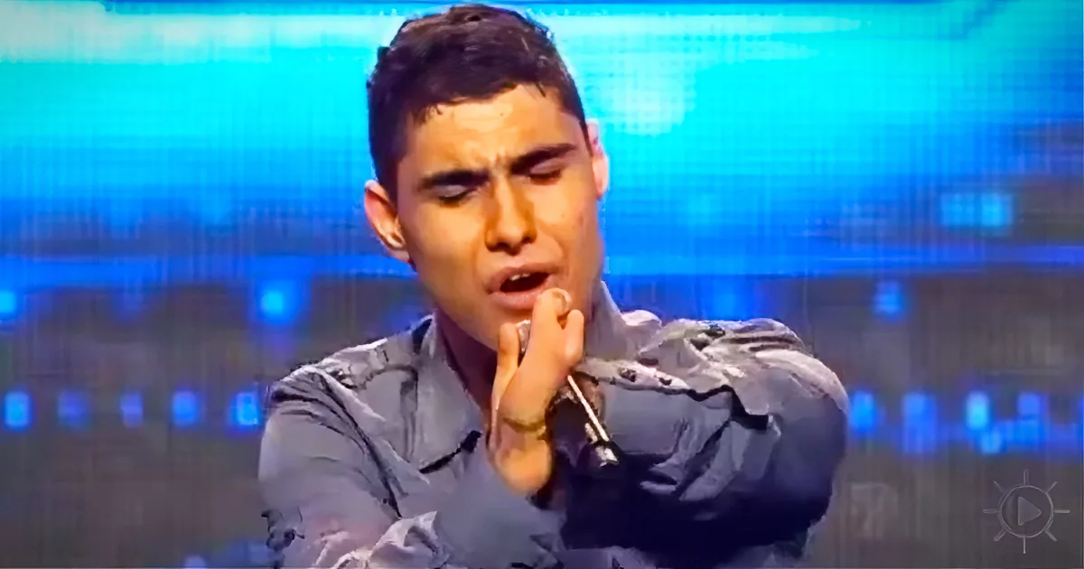 A person in a dark shirt gestures with their hand near their face against a blue-lit background.