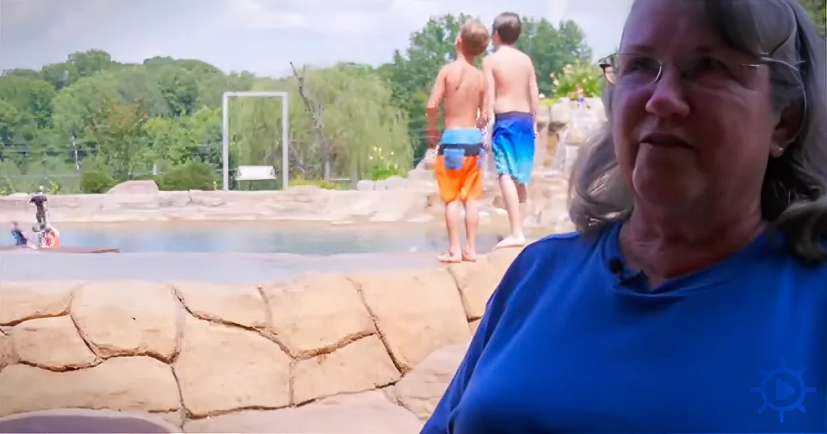 A woman in a blue shirt sits in front of a stone wall, with two boys in swim trunks walking away in the background.