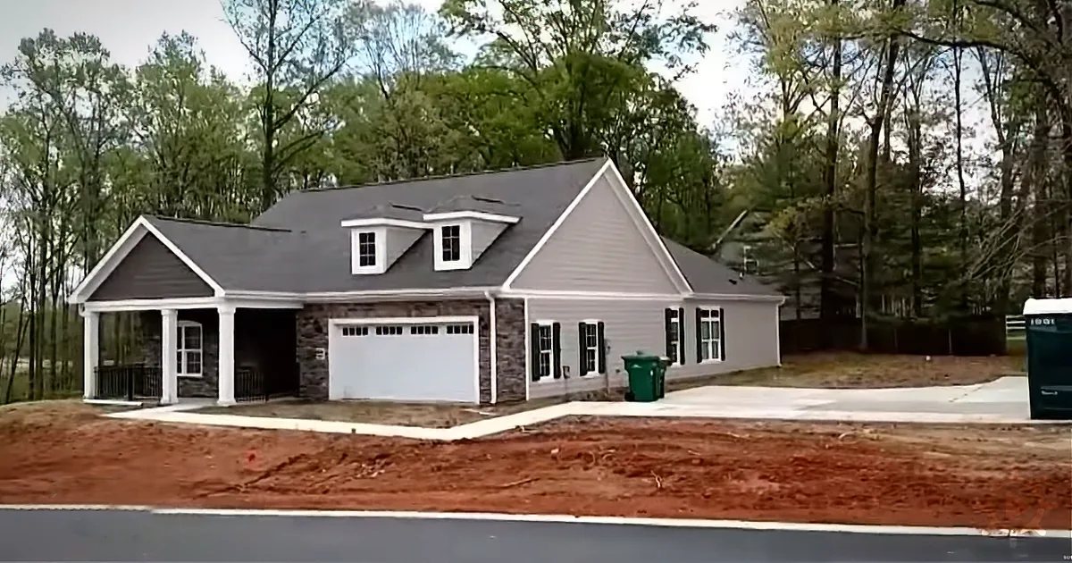 A newly built house with a white exterior, dark roof, and a two-car garage, surrounded by trees and dirt.