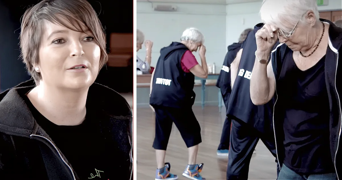 A group of elderly people in a gym are performing a synchronized exercise with their arms crossed in front of their chests.