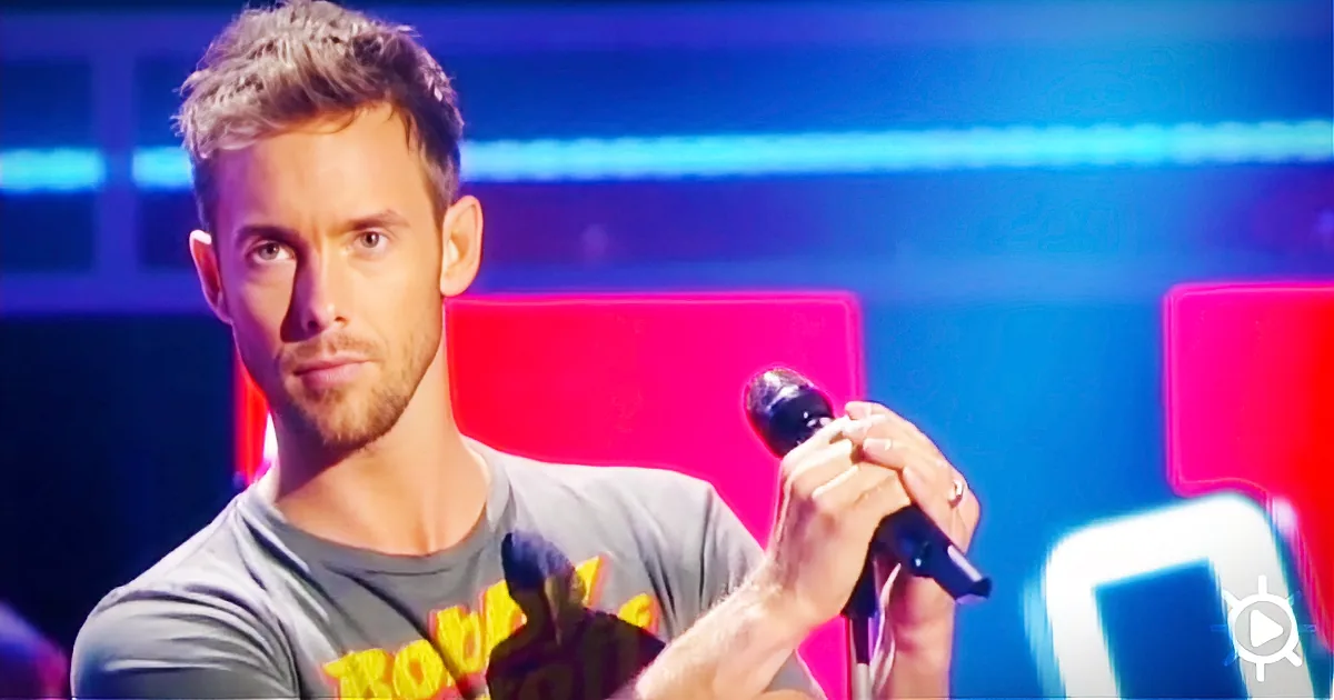 A man with short hair and a beard holds a microphone, wearing a gray t-shirt, against a colorful background.