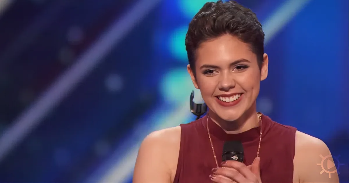 A smiling woman in a maroon dress holds a microphone, standing on a stage with a blue background.
