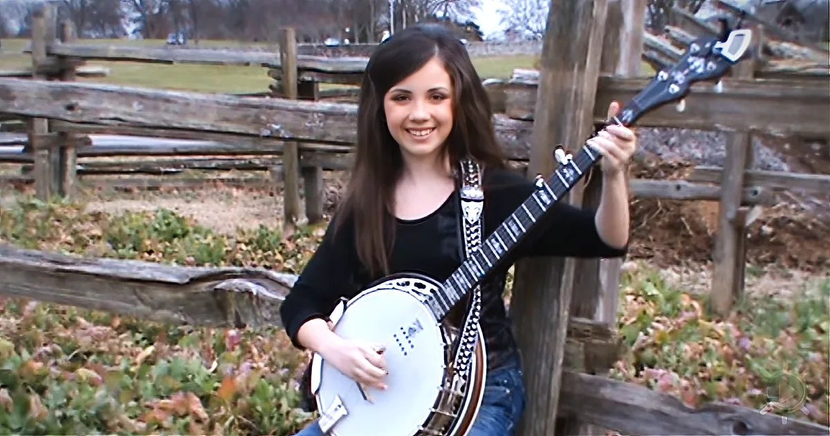 A girl with long brown hair smiles while holding a white banjo in front of a wooden fence.