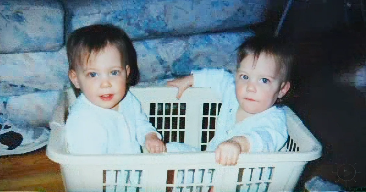 Two young children in a white laundry basket, looking serious, with a blue wall and shoes in the background.
