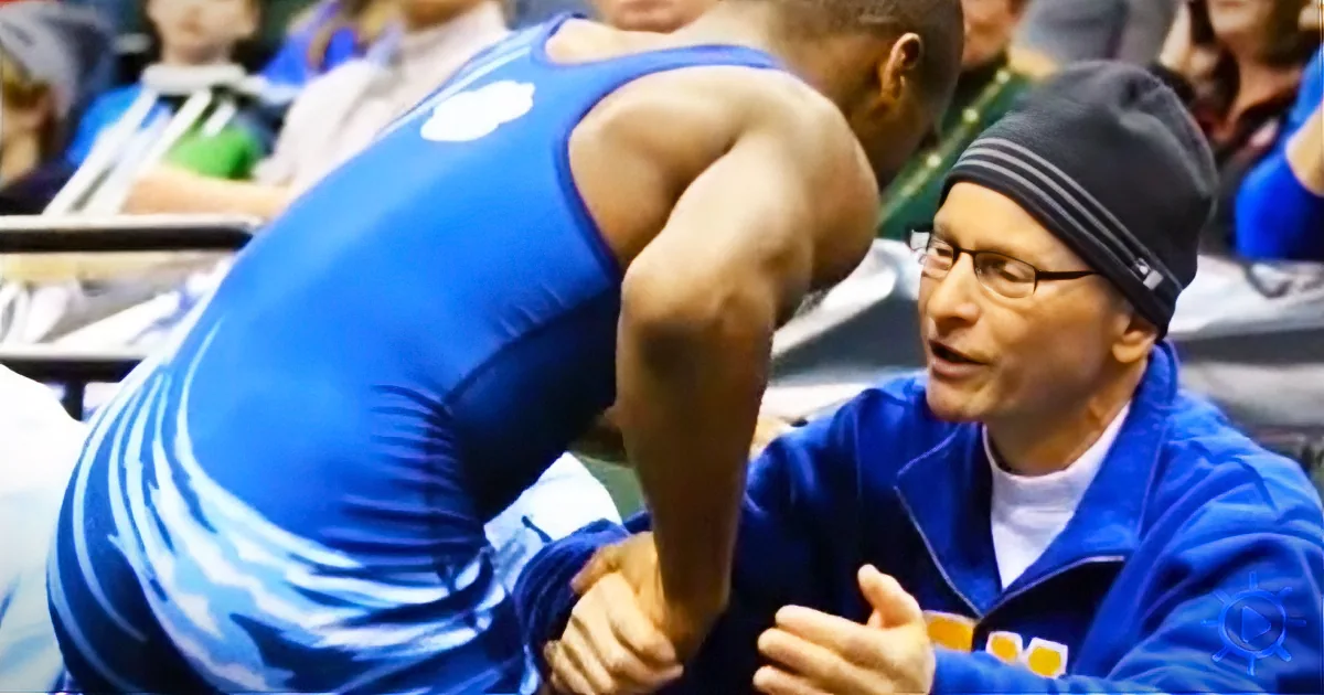 A wrestler in a blue singlet is being assisted by a coach in a blue jacket and beanie.