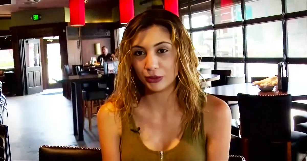A woman with long, wavy hair sits in a restaurant, wearing a sleeveless top, with a window and red lamps in the background.