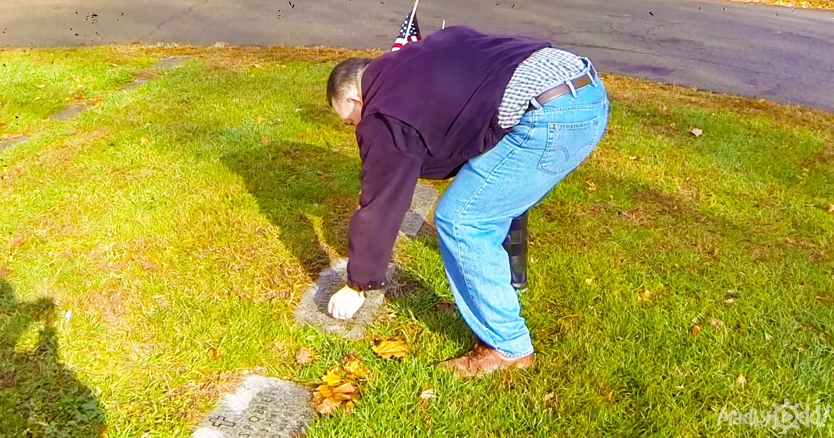 A man in jeans and a purple shirt bends over on grass, reaching towards a grave marker.