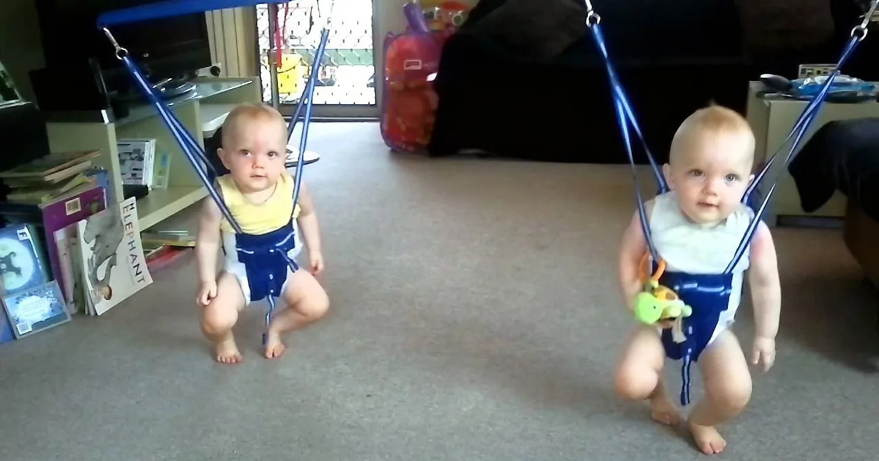 A baby wearing a blue and white harness is squatting and holding a green toy in a room with books and a suitcase.