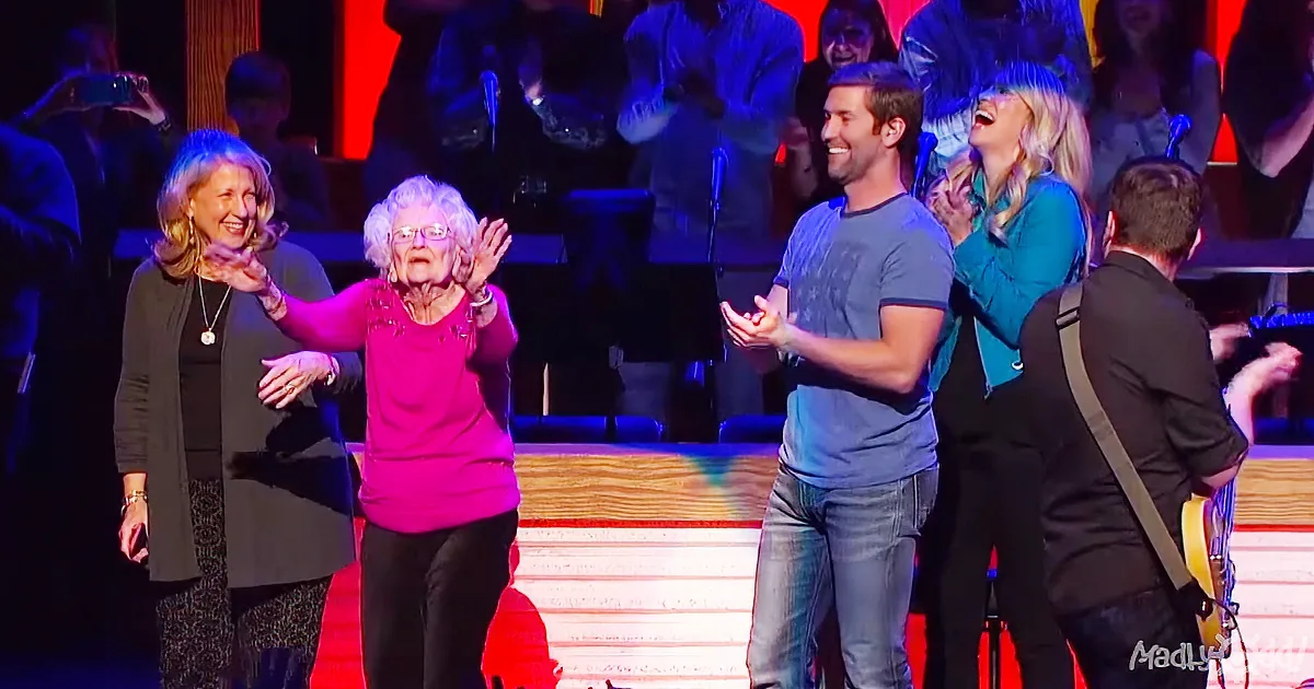 An elderly woman in a pink shirt is clapping on stage with others, surrounded by a cheering audience.