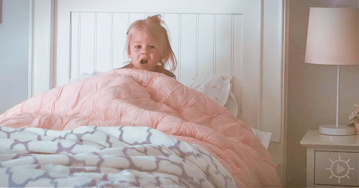 A child sits on a bed with a pink comforter, surrounded by pillows and a lamp on a nightstand.