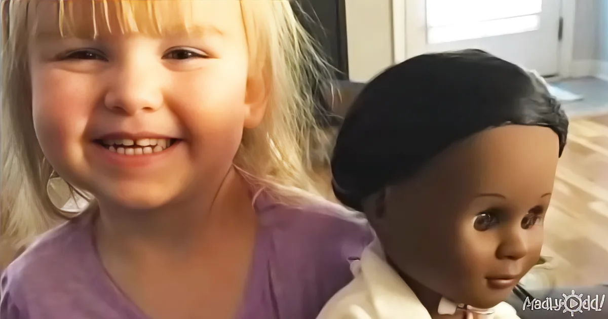 A smiling child with blonde hair and a purple shirt is holding a doll with dark hair and a white shirt.