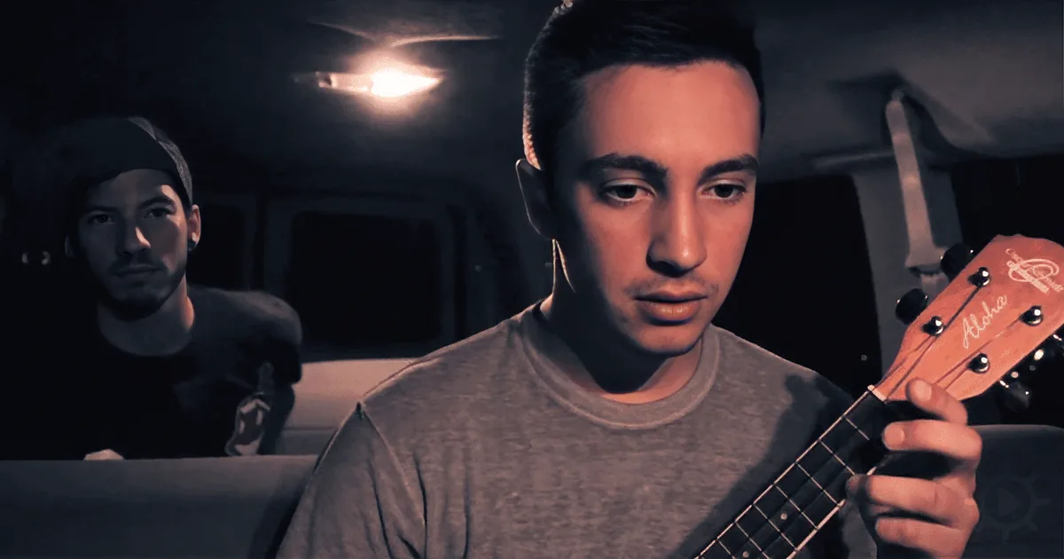 A man in a gray shirt holds a ukulele, looking down, with another person in the background.