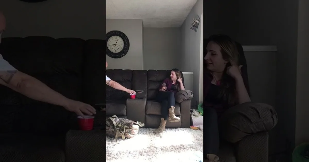 A woman sits on a couch, holding a phone, while another person sits nearby with a red cup.