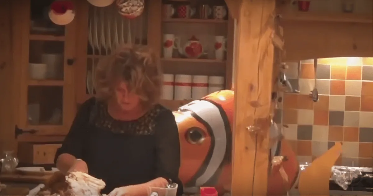 A person in a kitchen, wearing a clownfish costume, is holding something wrapped in foil.