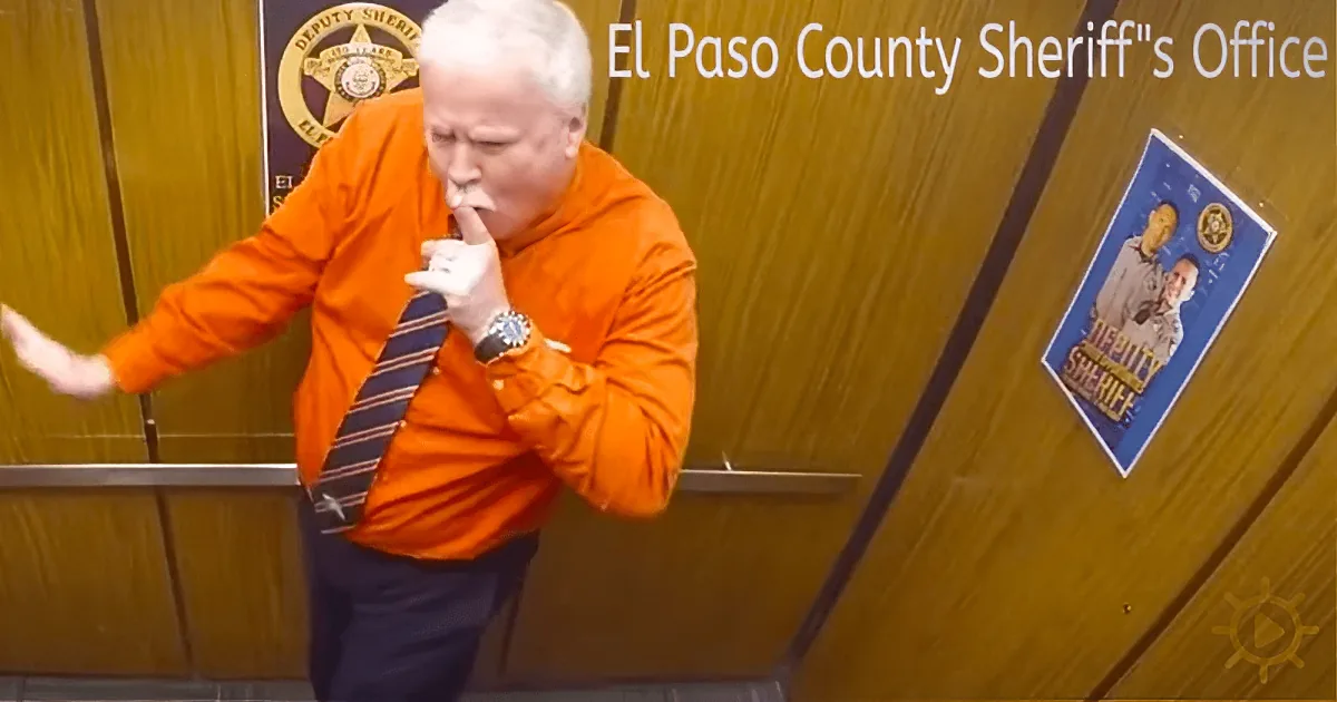 A man in an orange shirt and tie stands in front of a wooden door with "El Paso County Sheriff's Office" signage.
