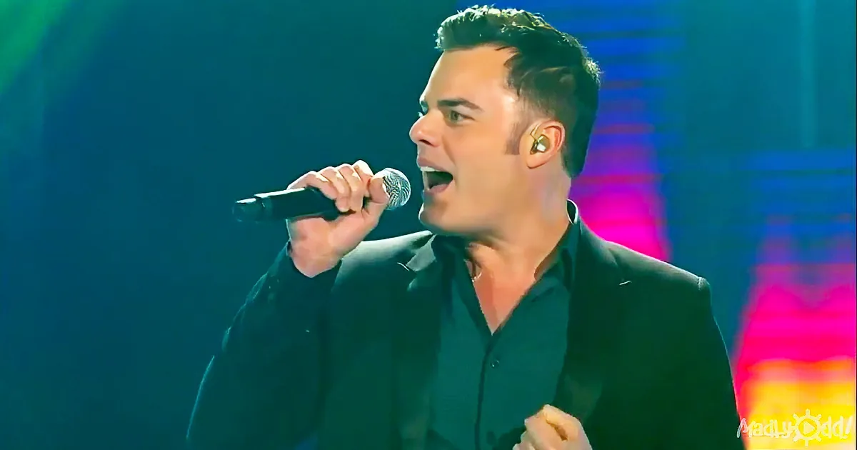 A man in a black suit sings passionately into a microphone on stage.