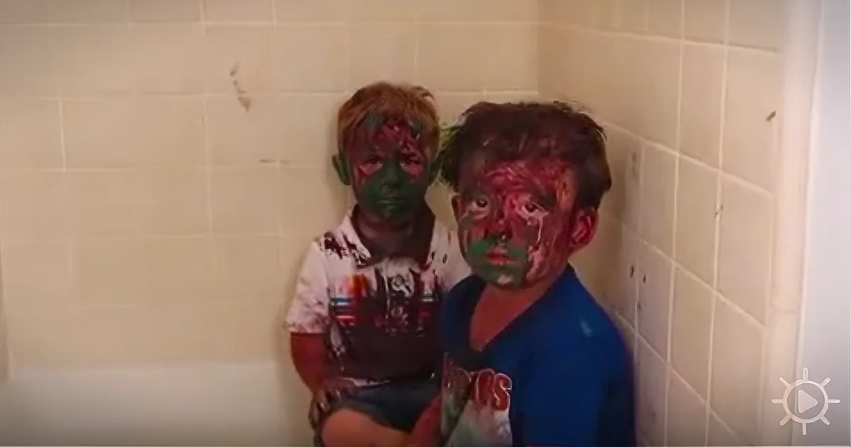 Two children with faces covered in paint stand against a tiled wall.