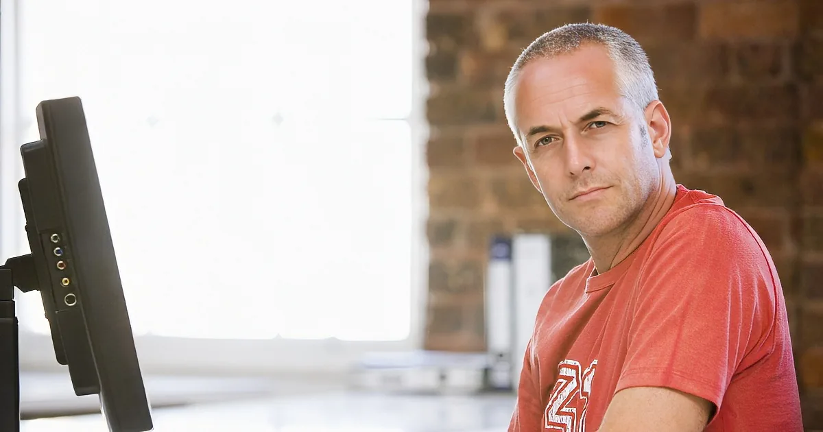 A man in a red shirt sits at a desk, typing on a keyboard and looking at the camera.