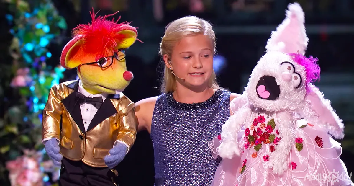 A young girl stands with two puppets, one in a gold suit and the other in a pink dress, on a stage.