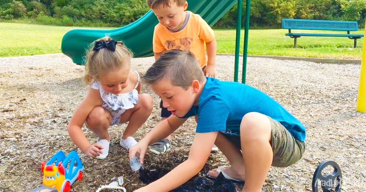 Children play with toys in a sandbox at a playground with a slide and bench in the background.