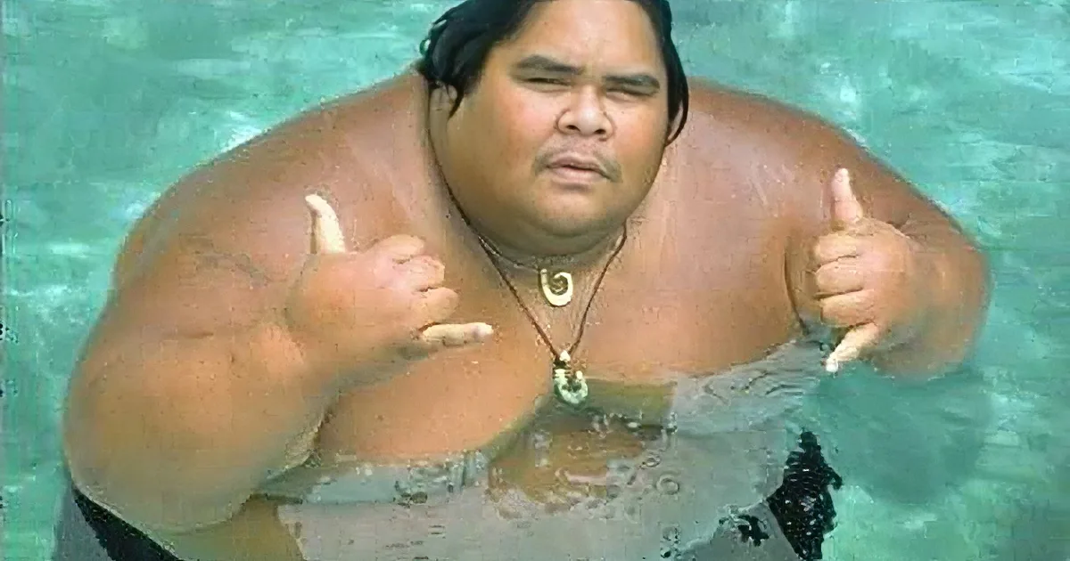 A man in a pool giving a shaka sign with both hands, wearing a necklace.