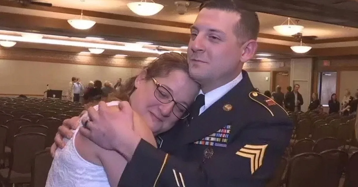 A soldier in uniform hugs a woman in a white top in a room filled with chairs and people.