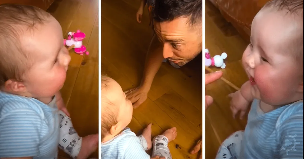 A baby in blue pajamas interacts with a toy and an adult on a wooden floor.