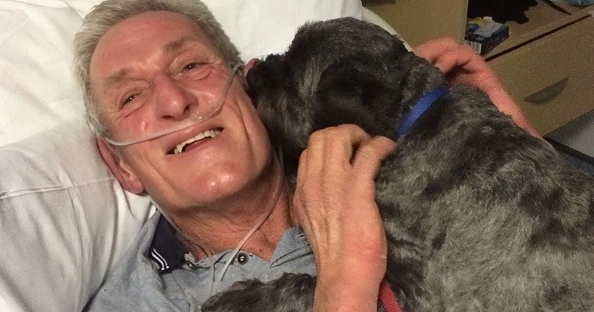 An elderly man with a tube in his mouth is lying in bed, cuddling a gray dog with a blue collar.