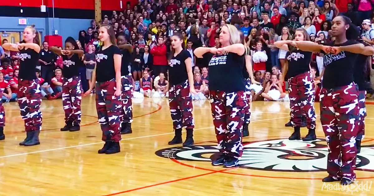A group of dancers in camouflage pants and black shirts perform on a basketball court in front of a large audience.