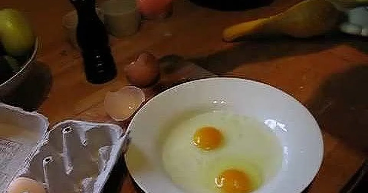 A cracked egg in a white bowl on a wooden table with egg carton and other ingredients nearby.