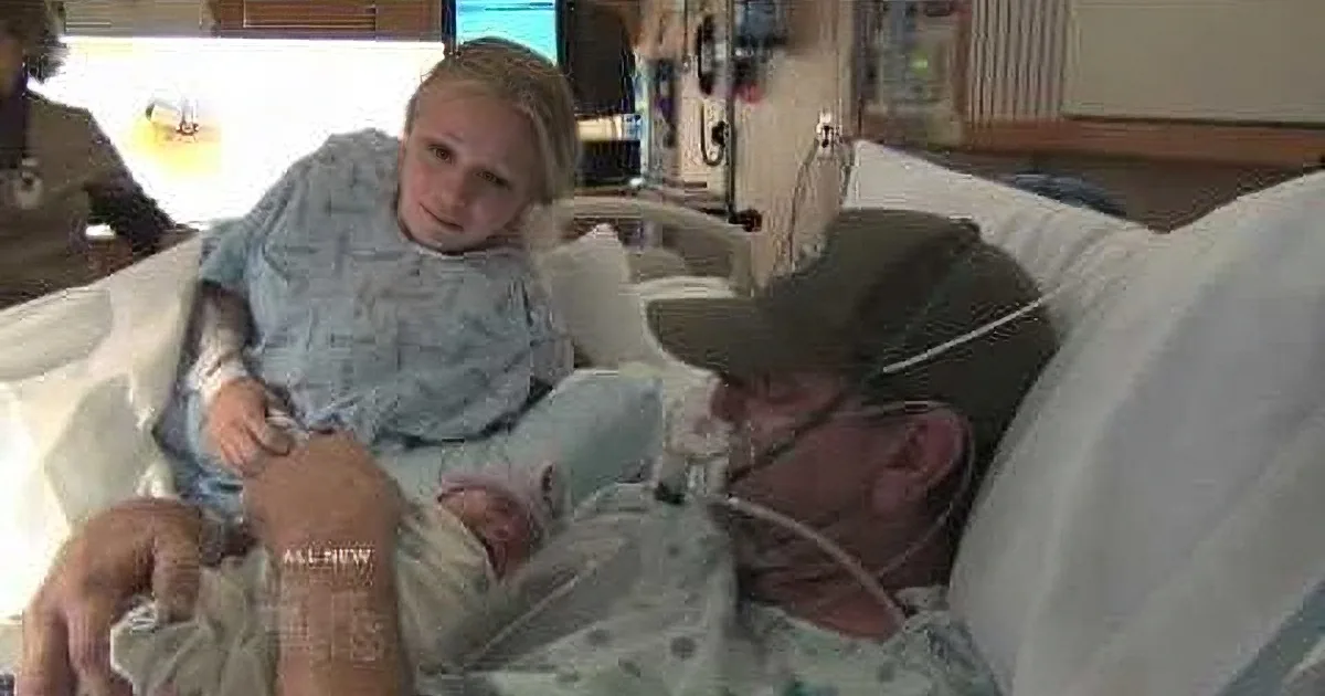 A woman in a hospital bed holds a newborn baby, with a man in a cap and oxygen mask lying beside her.