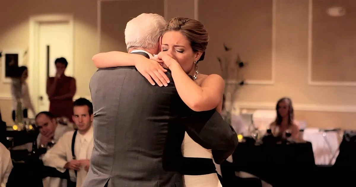 A woman in a white dress hugs a man in a suit at a formal event, with guests in the background.
