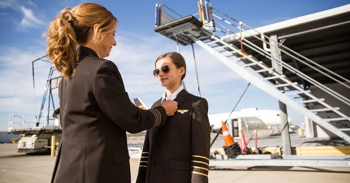 A woman in a pilot uniform adjusts a young boy's epaulets on an airport tarmac.