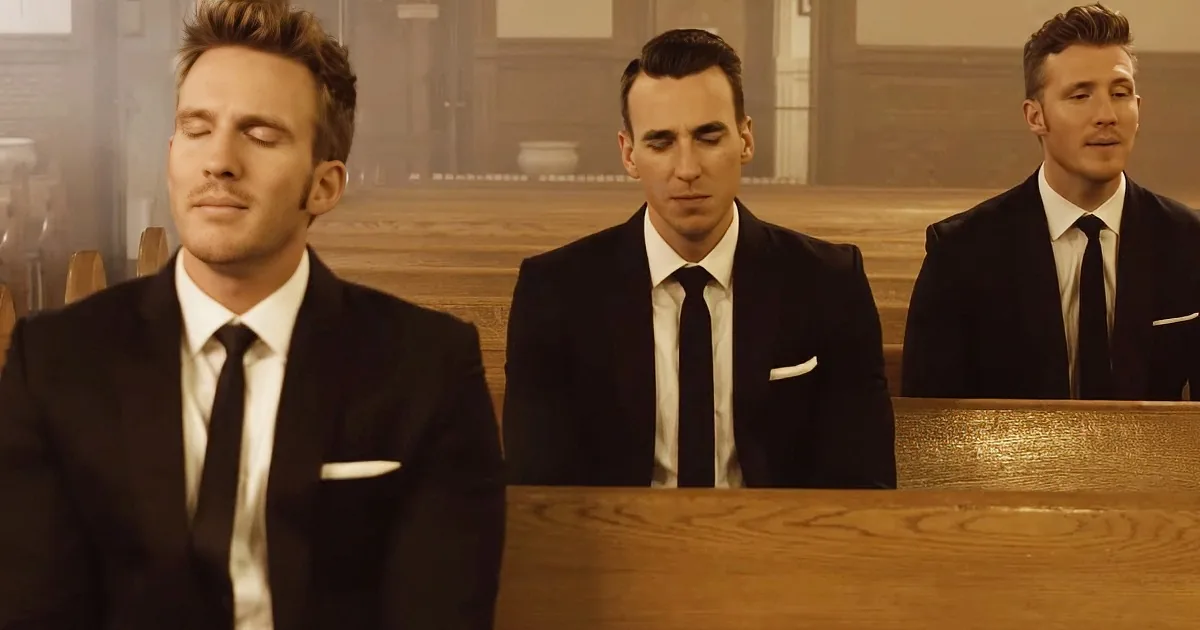 Three men in suits sit on wooden pews, eyes closed, in a church setting.